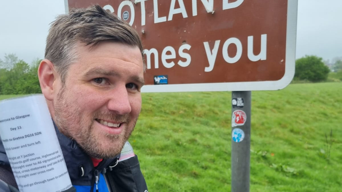 Andrew Williams stands next to a Welcome to Scotland sign