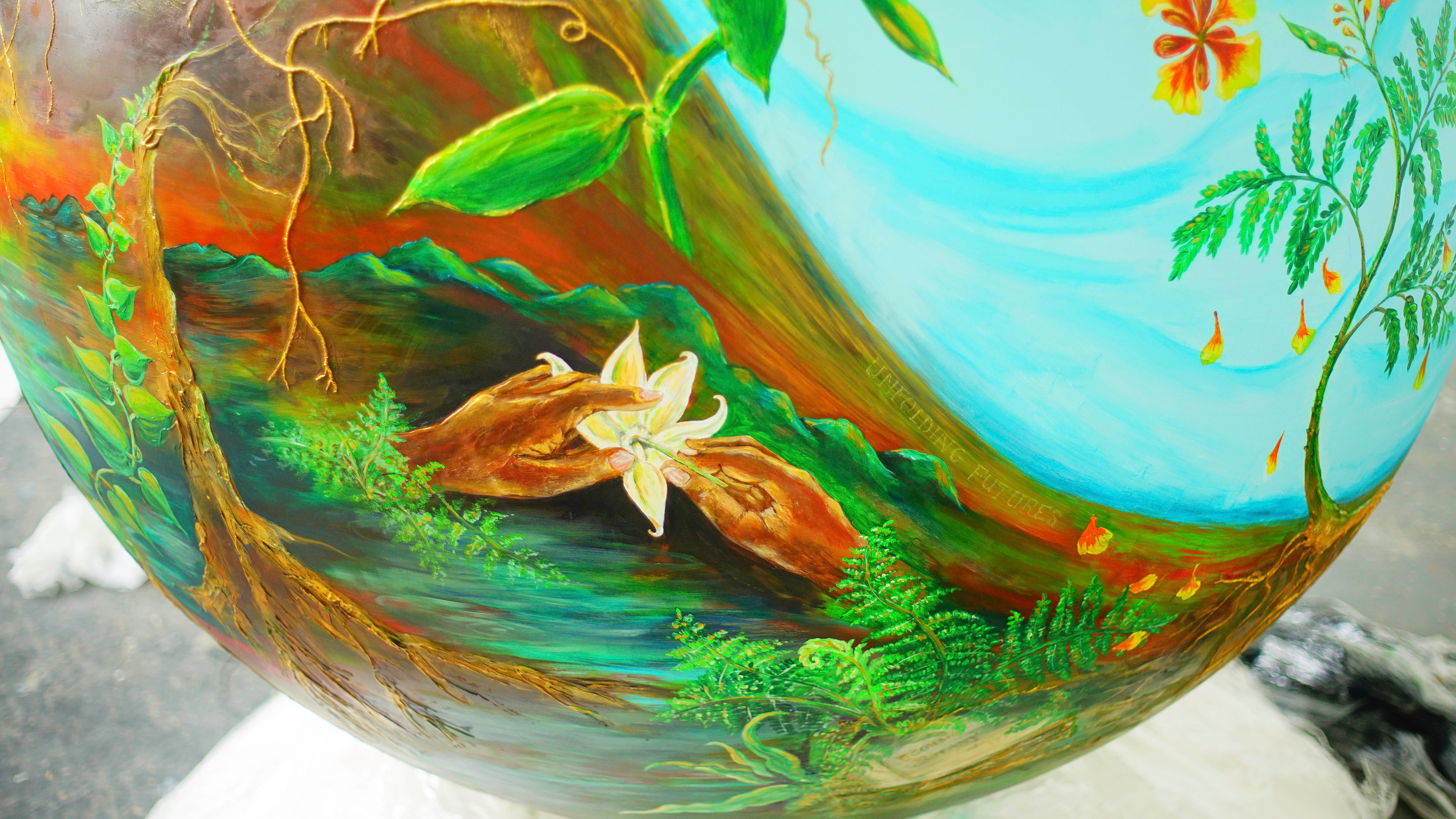 A close up of the globe painted by Carol Sorhaindo for Swansea. There are two hands holding a white flour, a landscape with plants, trees and mountains.