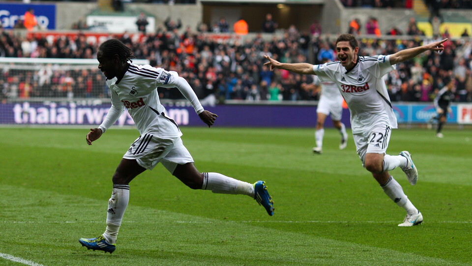 Two football players celebrate a goal in front of a cheering crowd.